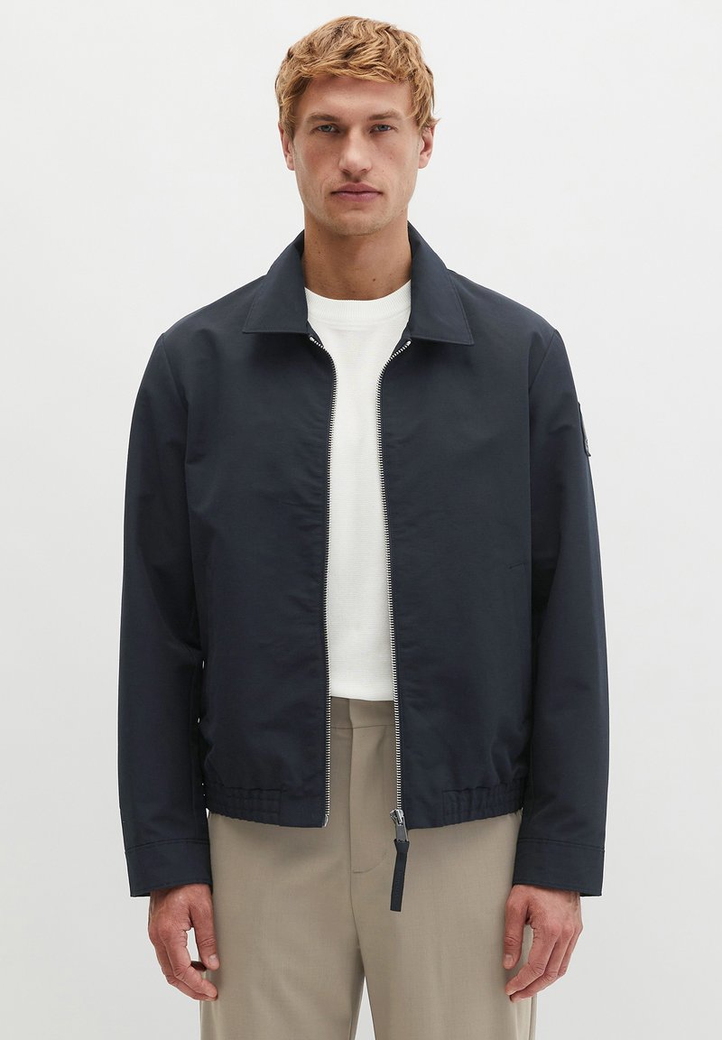 Blonder Mann trägt eine marineblaue Jacke mit Reißverschluss, einen weißen Pullover mit Rundhalsausschnitt und beige Hose und steht vor einem schlichten hellen Hintergrund.
