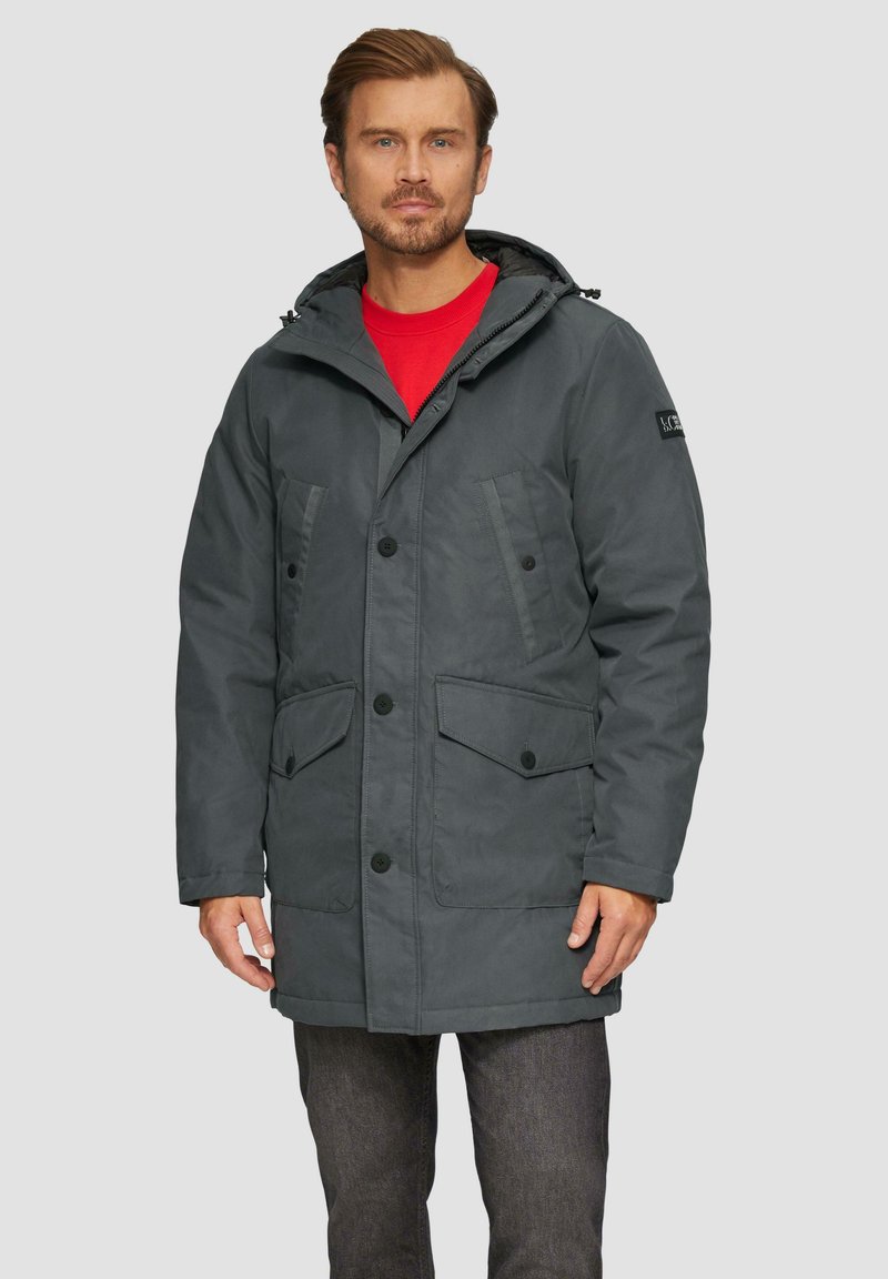 Parka grise avec une capuche, fermeture à boutons, poches avant et texture lisse, portée par-dessus une chemise rouge.