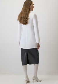 Garām piedurknēm balts tops ar taisnu piegriezumu un aizmugures šuvi, saskaņots ar tumšu ādas svārku, kuram ir centrāla šķēlums un bēšas zeķbikses.