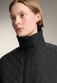 Giubbotto nero trapuntato con colletto alto. Presenta una superficie testurizzata con motivi a rombi e una chiusura a zip sul davanti.