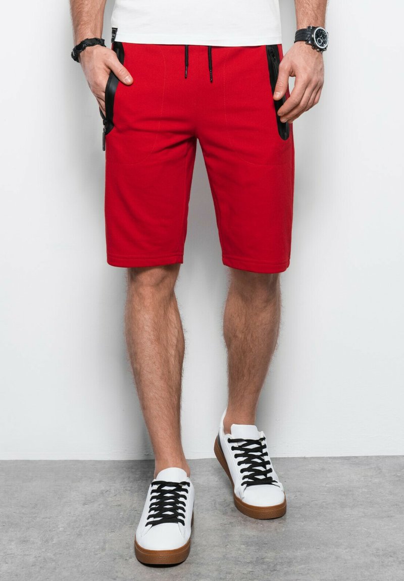 Shorts rouges avec des poches latérales zippées, fabriqués en tissu texturé. Associés à des baskets blanches avec une semelle marron et des lacets noirs.