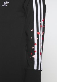 Czarna bluza z białymi paskami Adidas na rękawach. Lewy rękaw ozdobiony jest haftowanymi czerwonymi i różowymi sercami oraz małymi logotypami. Miękka tekstura materiału.