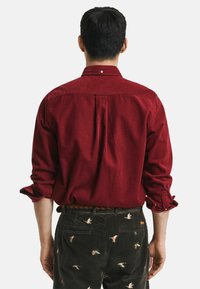 Rode corduroy overhemd met een kraag, zichtbare rugplooien en button cuffs, gedragen met olijfgroene broek met geborduurde eenden.