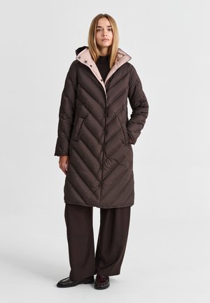 Femme portant un long manteau matelassé marron foncé avec une doublure de capuche rose clair, un haut noir, un pantalon marron ample et des chaussures noires, debout sur un fond uni.