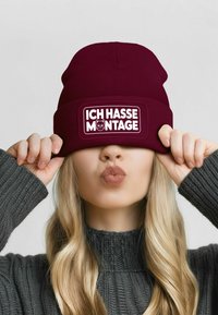 Maroon knit beanie with a rectangular white patch displaying the text "ICH HASSE MONTAGE" and a cat graphic. Soft texture, snug fit.