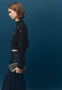 Maglione nero corto con embellishments argentati rotondi, abbinato a pantaloni in denim scuro e una pochette argentata texturizzata. Sfondo blu navy.