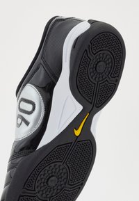 Crne atletske cipele sa sjajnim gornjim dijelom, koje imaju veliki bijeli okrugli simbol s brojem "90" u sivoj boji i žutom Nike logotipu na potplatu.