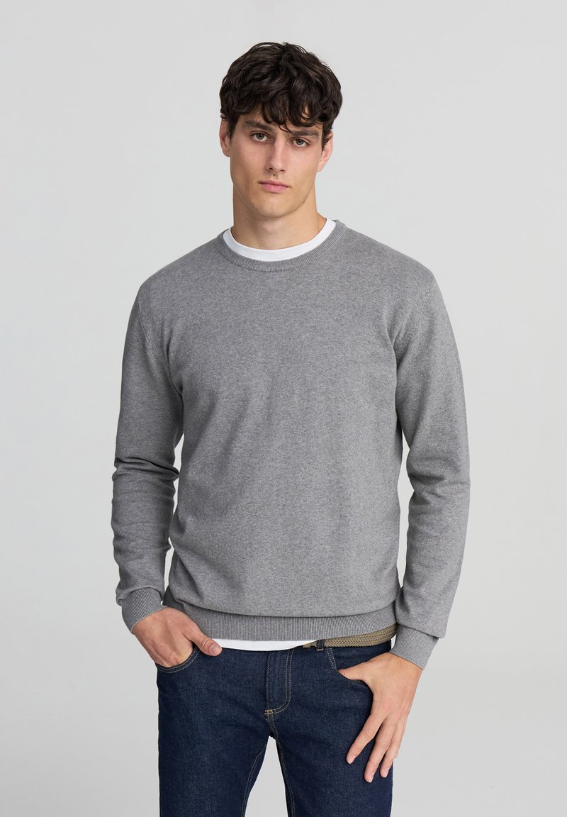 Felpa grigia a girocollo in morbido tessuto con maniche lunghe e polsini a costine. Indossata sopra una t-shirt bianca, abbinata a jeans blu scuro.