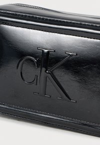 Črna usnjena kozmetična torbica z sijajnim finišem, z embosiranim logotipom "CK", zadrgo in gladko teksturo.