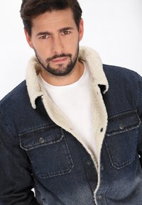 Farkkutakki tummansinisellä sävyllä, jossa on vaaleanbeige fleecekaulus, kaksi rintataskua ja nappikiinnitys. Alla on yksinkertainen valkoinen paita.