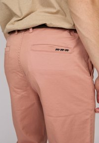 Pantalons rose clair avec une texture lisse, comportant une poche arrière et des coutures avec un logo d'animal noir. Design décontracté avec une coupe ajustée.