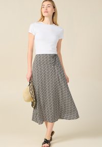 Jupe maxi noire avec un motif floral crème, tissu léger, design fluide, associée à une chemise blanche ajustée et des sandales noires.