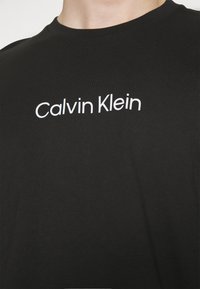 T-shirt en coton noir avec un logo "Calvin Klein" blanc. Design col rond, texture lisse, avec des manches courtes et une coupe ajustée.