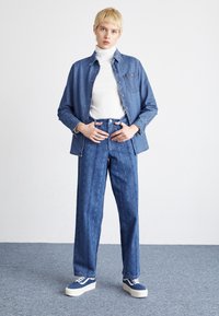 Veste en jean avec des boutons sur un col roulé blanc, associée à un jean large à motifs bleus et des baskets bleues avec des semelles blanches.