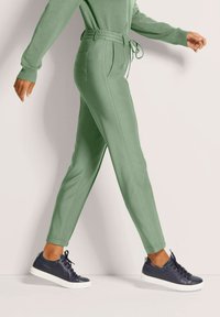 Persoon die loopt in lichtgroene broek met een strikriem en zwarte sneakers met witte zolen, op een neutrale achtergrond.