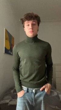 Young man with curly brown hair wearing a green turtleneck and light blue jeans, standing indoors with hands in pockets.