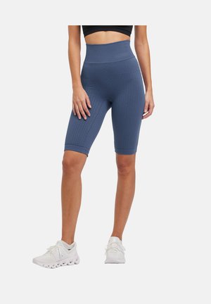 Frau trägt hochtaillierte, strukturierte navyblaue Radlerhosen und weiße Sportschuhe, steht vor einem schlichten weißen Hintergrund.