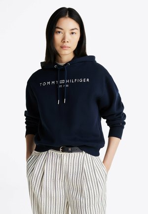 Felpa navy con cappuccio e cordino, con logo bianco "TOMMY HILFIGER" e testo "EST. 1985". Modello abbinato a pantaloni a righe e una cintura discreta.