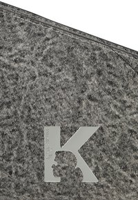 Γκρίζο υφασμάτινο υλικό με υφή και σχέδιο, που περιλαμβάνει ένα προβεβλημένο ασημένιο λογότυπο "Κ". Ο σχεδιασμός περιλαμβάνει διακριτές παραλλαγές στη σκιά και την υφή.