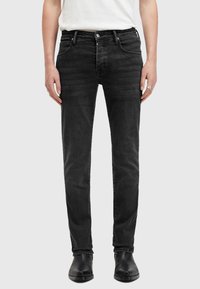 Černé džínové kalhoty s úzkým střihem, vybavené pěti kapsami, zipem a jemnými detaily v podobě šisování, doplněné černými kotníkovými botami.