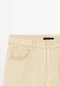 Pantalon beige taille haute avec fermeture par bouton, passants de ceinture et détail de poche avant, affichant l’étiquette de la marque Massimo Dutti.
