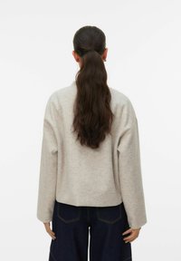 Femme aux longs cheveux foncés attachés en queue de cheval basse, portant un pull beige et un jean bleu foncé, vue de dos sur un fond blanc.