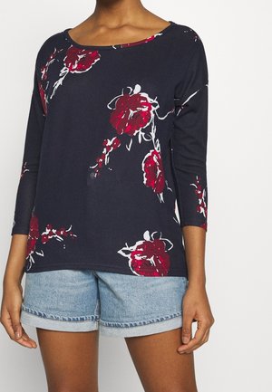 Personne portant un haut à manches longues bleu marine avec un imprimé floral rouge et blanc et un short en jean bleu clair sur fond blanc uni.