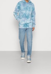Light blue tie-dye hoodie with front pocket, drawstring hood, and white logo on the left chest. Paired with light blue jeans and sneakers.