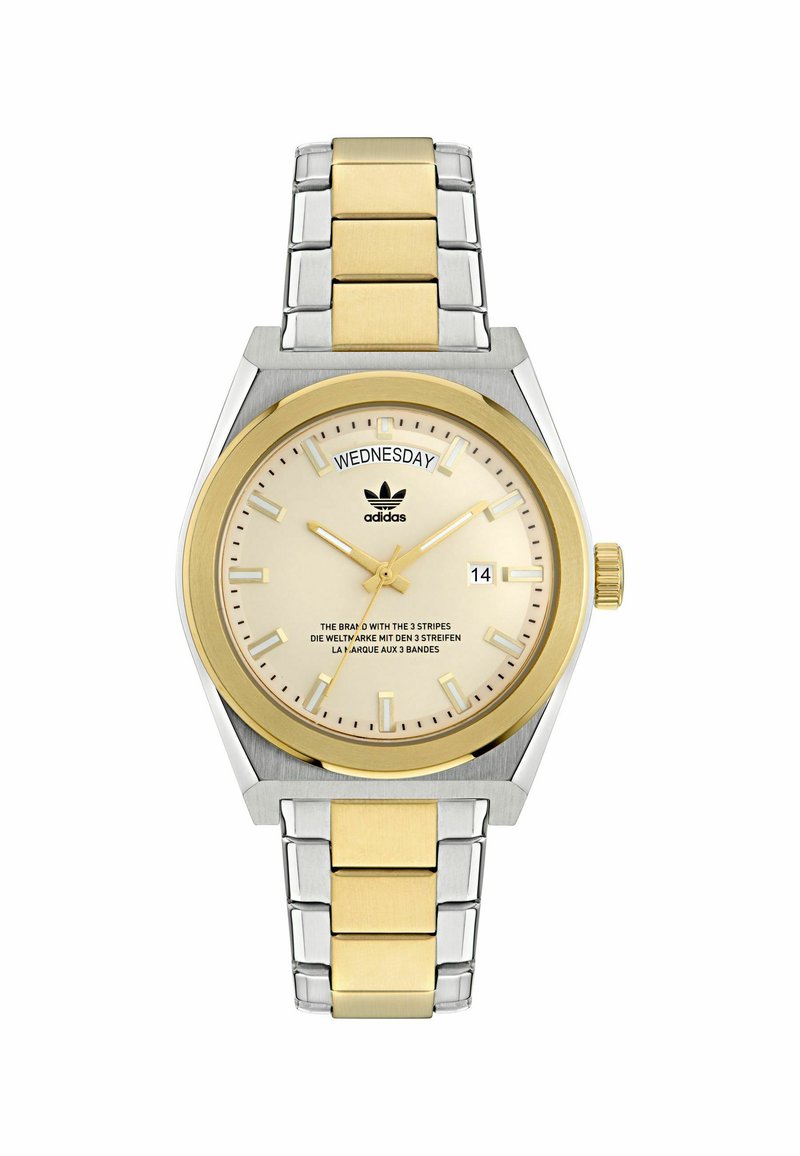 Kullatud ja hõbedane metallkell ümmarguse korpusega, kreemikaskellakuva, kuupäevanäidiku, tunni markerite ja Adidas logo; omaduseks on kahetooniline metallist rihm.
