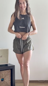 Femme souriante ajuste le cordon de serrage de son short vert olive, portant un débardeur noir court, debout pieds nus à l’intérieur près d’un haut-parleur et d’un panier en osier.