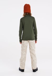 Top verde de manga larga con un diseño ajustado, combinado con pantalones beige estampados. El conjunto se completa con un gorro de punto naranja texturizado.