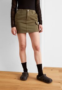 Olivenfarge cargo skjørt med frontlommer, tettsittende design, og hem over midt låret. Kombinert med svarte sokker og svarte tresko.
