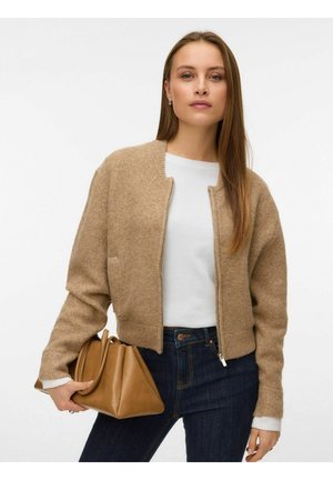 Gilet en tricot brun avec un col rond, des poignets côtelés et une fermeture éclair à l'avant, associé à une chemise blanche et un jean en denim foncé, portant un sac brun.