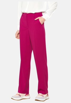 Femme portant un pantalon droit d’un rose magenta vif avec des poches latérales, associé à des baskets blanches et une chemise blanche à manches longues.