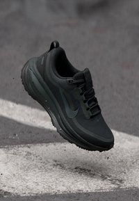 Scarpa da corsa nera Nike in aria sopra l'asfalto con strisce bianche sulla strada, piccoli detriti che cadono attorno alla suola.