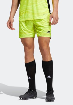 Personne portant un short de soccer Adidas jaune néon, un maillot assorti, des chaussettes montantes noires Adidas et des crampons noirs, debout sur un sol clair.