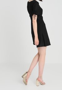 Robe noire courte avec des manches bouffantes et des détails à cordon, associée à des sandales à talons hauts métalliques dorées avec une bride de cheville.