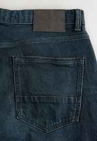 Jeans din denim de culoare albastră închisă, având un singur buzunar pe partea din spate, etichetă din piele la talie și detalii de cusături vizibile.