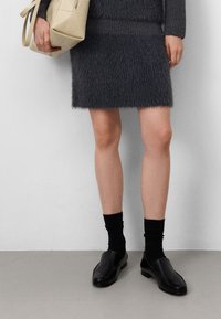Grijze nepbont mini-rok, geribbeld truitje in de taille, gecombineerd met zwarte sokken en instappers van leer. Beige tas in de rechterhand vastgehouden.