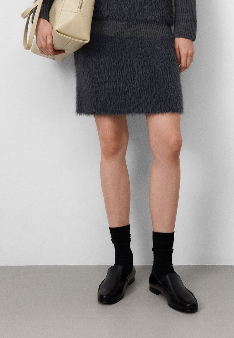 Grauer Kunstpelz-Minirock, gerippter Pullover an der Taille, kombiniert mit schwarzen Socken und lässigen Lederschuhen. Beigefarbenes Täschchen in der rechten Hand gehalten.