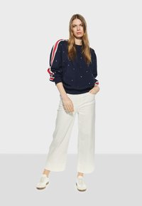 Marinblå sweatshirt med vita prickar och röda randiga detaljer på ärmarna, matchad med vida vita byxor och vita sneakers.