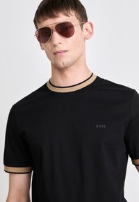 Schwarzes Rundhalsausschnitt-T-Shirt mit kontrastierendem, beige farbenem Besatz am Kragen und an den Ärmeln, das ein dezentes Markenlogo auf der Brust trägt. Triviale Textur.