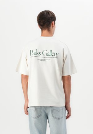 Person wearing a white oversized T-shirt with "Parks Gallery" text on the back, paired with light blue jeans, standing against a plain background.