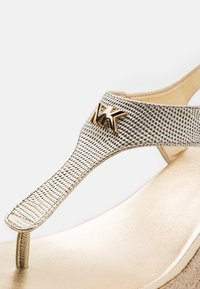 Lanière de sandale tongs texturée dorée avec un charme logo MK métallique, attachée à une semelle intérieure dorée et une base de talon compensé tissée.