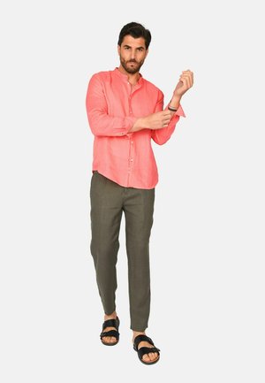 Uomo che sistema la manica indossando una camicia corallo con bottoni, pantaloni oliva e sandali neri, in piedi contro uno sfondo chiaro e semplice.