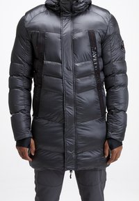 Man in een lange zwarte pufferjack met capuchon, ritssluiting aan de voorkant en zwarte panelen met rode stiksels op de borst, gecombineerd met grijze broek.