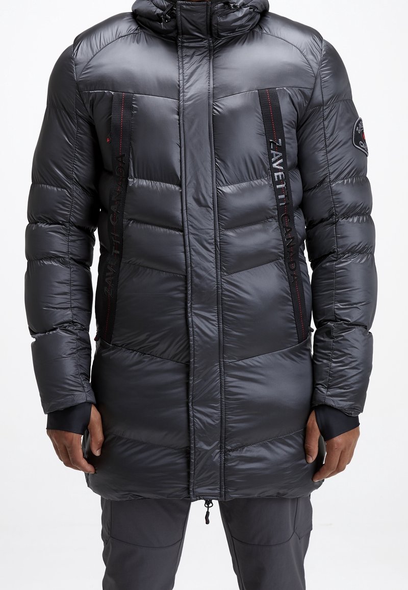 Man in een lange zwarte pufferjack met capuchon, ritssluiting aan de voorkant en zwarte panelen met rode stiksels op de borst, gecombineerd met grijze broek.