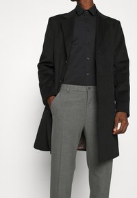 Cappotto lungo in lana nera, pantaloni grigi su misura e camicia nera con bottoni. Caratteristiche includono taglio dritto e leggera texture.