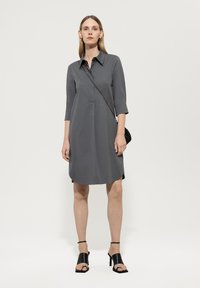 Graues Hemdkleid mit Kragen, dreiviertel Ärmeln und gerader Silhouette. Das Model hält eine kleine schwarze Tasche.