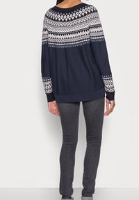 Maglione lavorato a maglia blu con un motivo geometrico bianco e rosa, caratterizzato da un colletto rotondo e maniche lunghe, abbinato a jeans grigi scuri attillati.